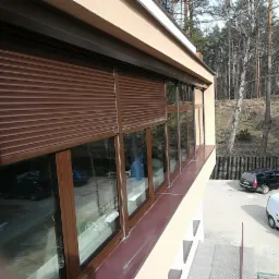 Brązowe rolety zewnętrzne zamontowane na oknach budynku, widok z zewnątrz, w tle las i zaparkowane samochody.