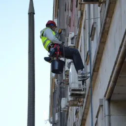 Pracownik na wysokości w uprzęży, z hełmem i linami, przy ścianie budynku z licznymi klimatyzatorami, wykonuje prace na wysokości.