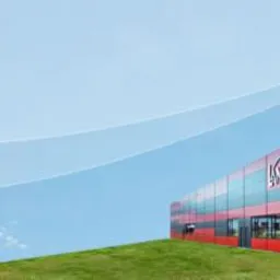 Budynek firmy Lange Kala w czerwono-szarej kolorystyce z napisem 'Ideen für's Dach' na tle błękitnego nieba i zielonego wzgórza.