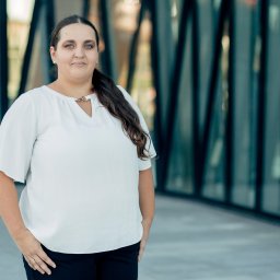 Poniewierka Radcowie Prawni spółka komandytowa - Portret kobiety w białej bluzce na tle szklanej fasady budynku. Uśmiech, długie włosy, delikatna biżuteria. Zdjęcie biznesowe, prawdopodobnie prawnik lub radca prawny.