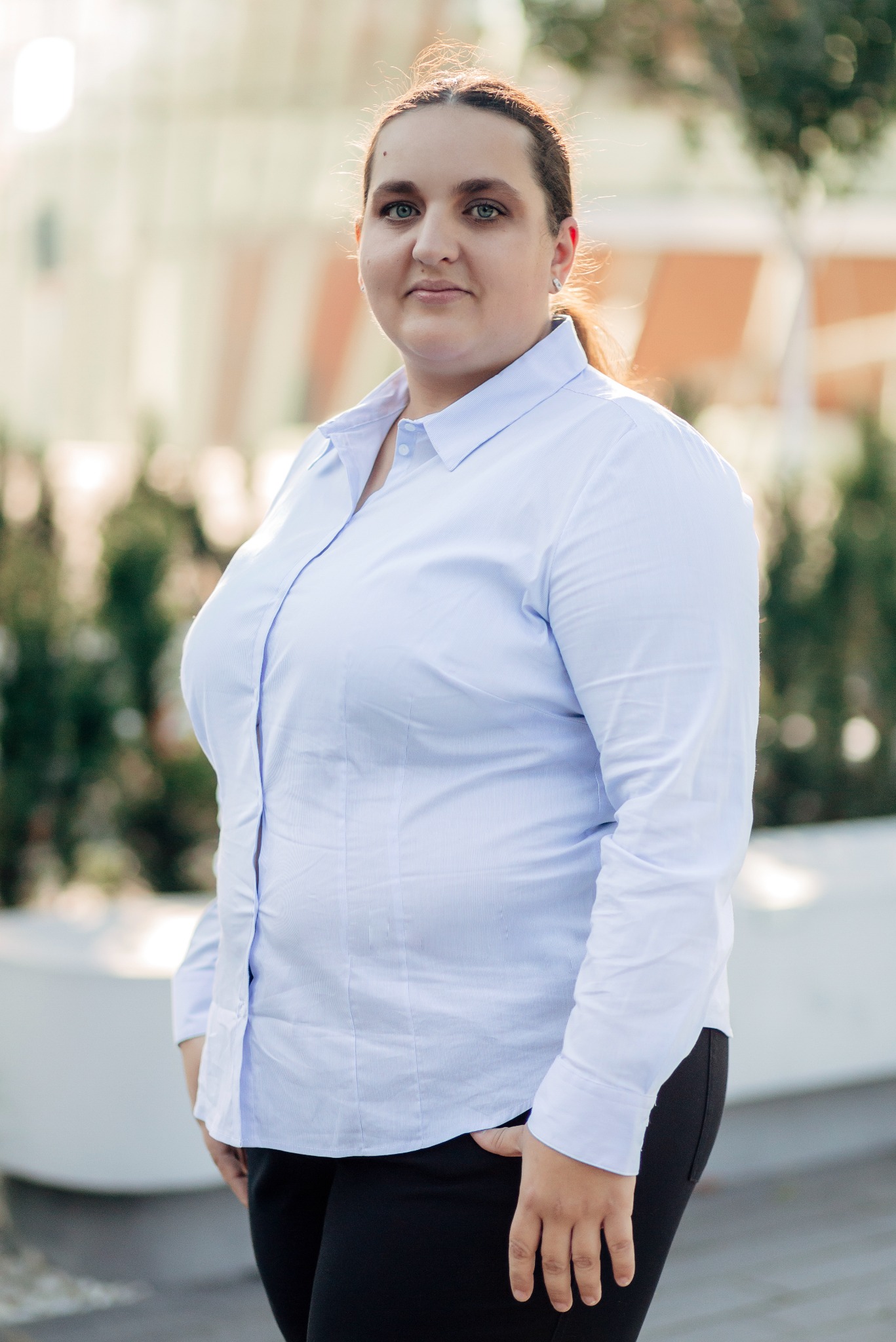 Portret kobiety w błękitnej koszuli na tle miejskiej zieleni i budynku. Uśmiech, profesjonalny wygląd, naturalne światło. Kadrowanie od kolan w górę.