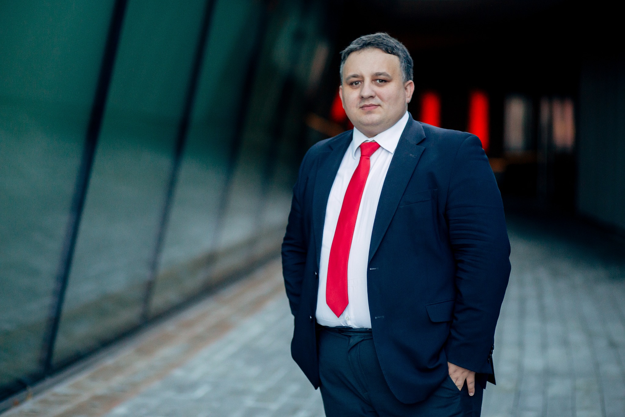 Portret mężczyzny w garniturze z czerwoną kravatą, stojącego na tle nowoczesnej architektury. Ujęcie od pasa w górę, ręce w kieszeniach, powaga w spojrzeniu.
