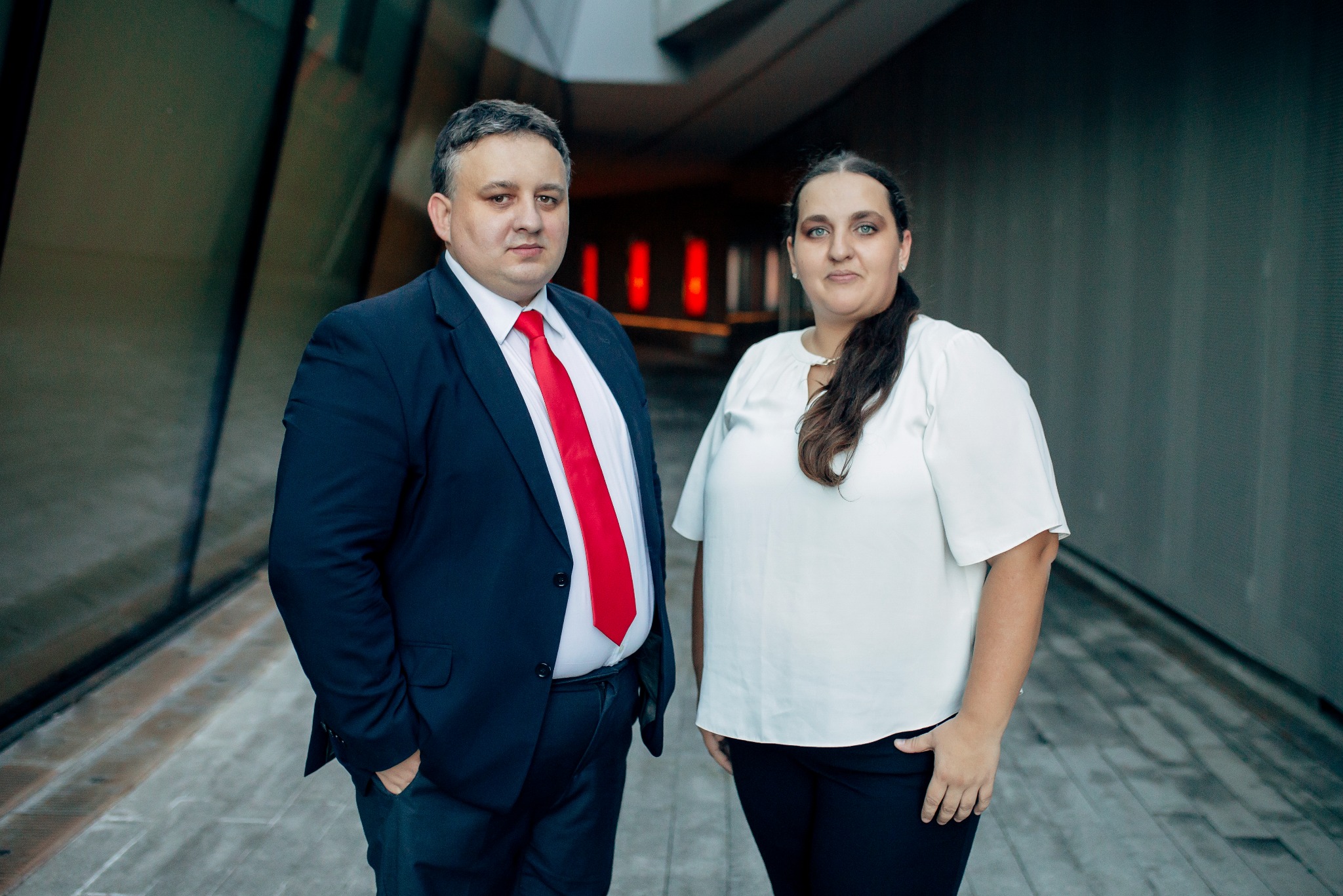 Portret dwojga prawników w eleganckich strojach na tle nowoczesnej architektury. Mężczyzna w garniturze z czerwoną kravatą, kobieta w białej bluzce. Stonowana kolorystyka.