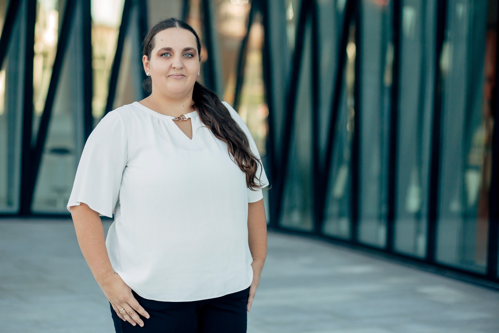 Portret kobiety w białej bluzce na tle szklanej fasady budynku. Uśmiech, długie włosy, delikatna biżuteria. Zdjęcie biznesowe, prawdopodobnie prawnik lub radca prawny.