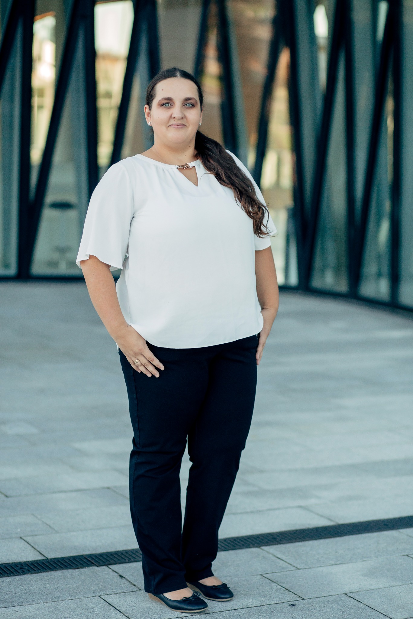 Portret kobiety w białej bluzce i czarnych spodniach na tle nowoczesnego budynku z przeszkloną fasadą. Uśmiechnięta, z rękami w kieszeniach, pozuje do zdjęcia.