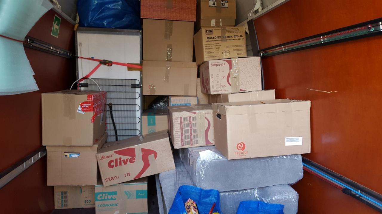 Kartonowe pudła i meble zabezpieczone pasami wewnątrz pojazdu transportowego. Widoczna lodówka i torby transportowe. Pomarańczowe ściany pojazdu.