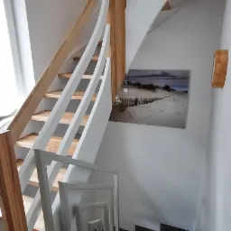 Spiralne schody z jasnego drewna i białymi balustradami, ciemne stopnie, na ścianie obraz z motywem plaży, widok z góry.