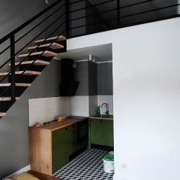 Lisowski - Stolarnia Paweł Lisowski - Nowoczesna kuchnia w aneksie z zielonymi szafkami, drewnianym blatem i czarno-białą podłogą w geometryczny wzór, widoczna spod drewnianych schodów z czarną balustradą prowadzących na antresolę.