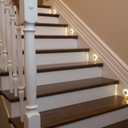 Lisowski - Stolarnia Paweł Lisowski - Klasyczne schody z ciemnego drewna i białymi podstopnicami, oświetlone punktowymi lampkami LED w ścianie, biała balustrada z toczonymi tralkami.