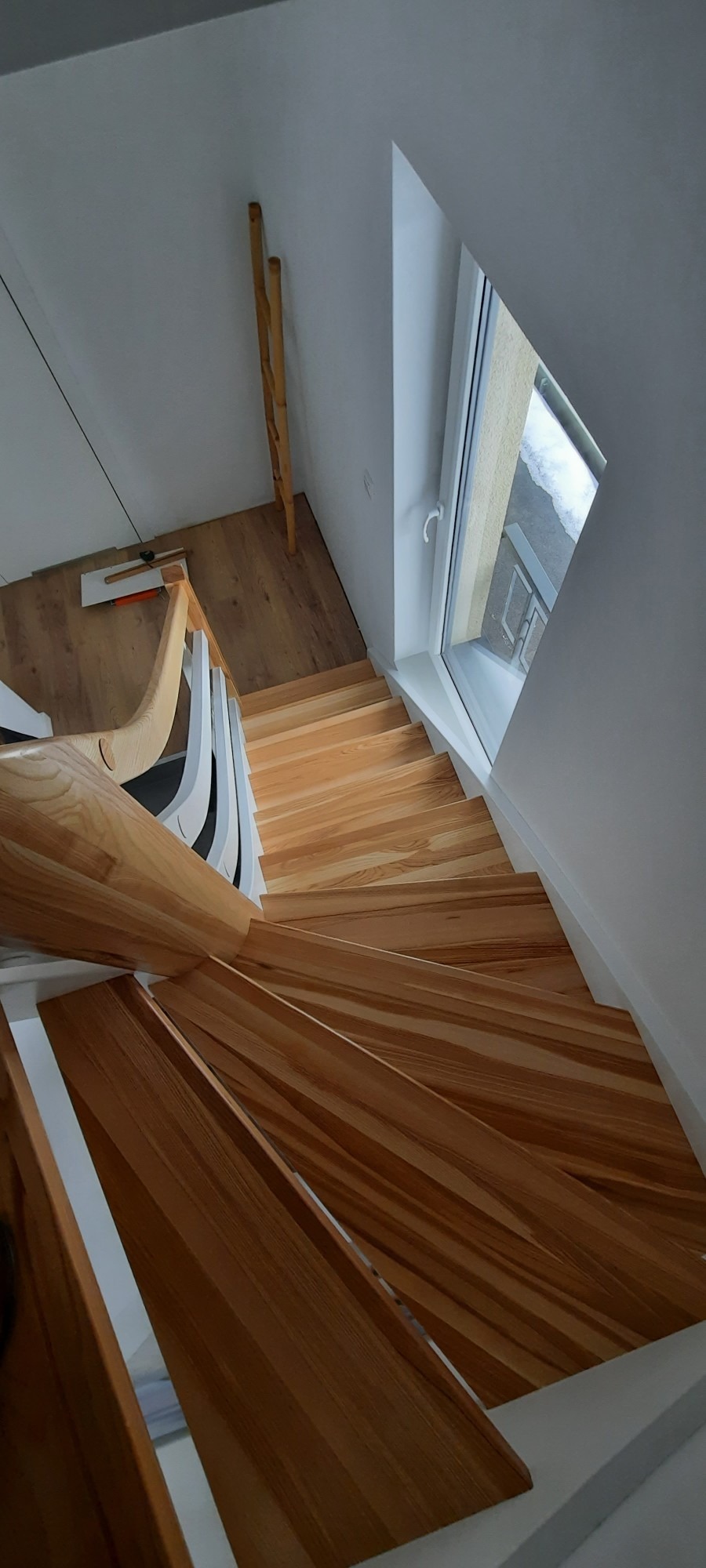 Spiralne schody z jasnego drewna z białymi balustradami, widok z góry na stopnie, w tle okno dachowe i drewniane podpory.