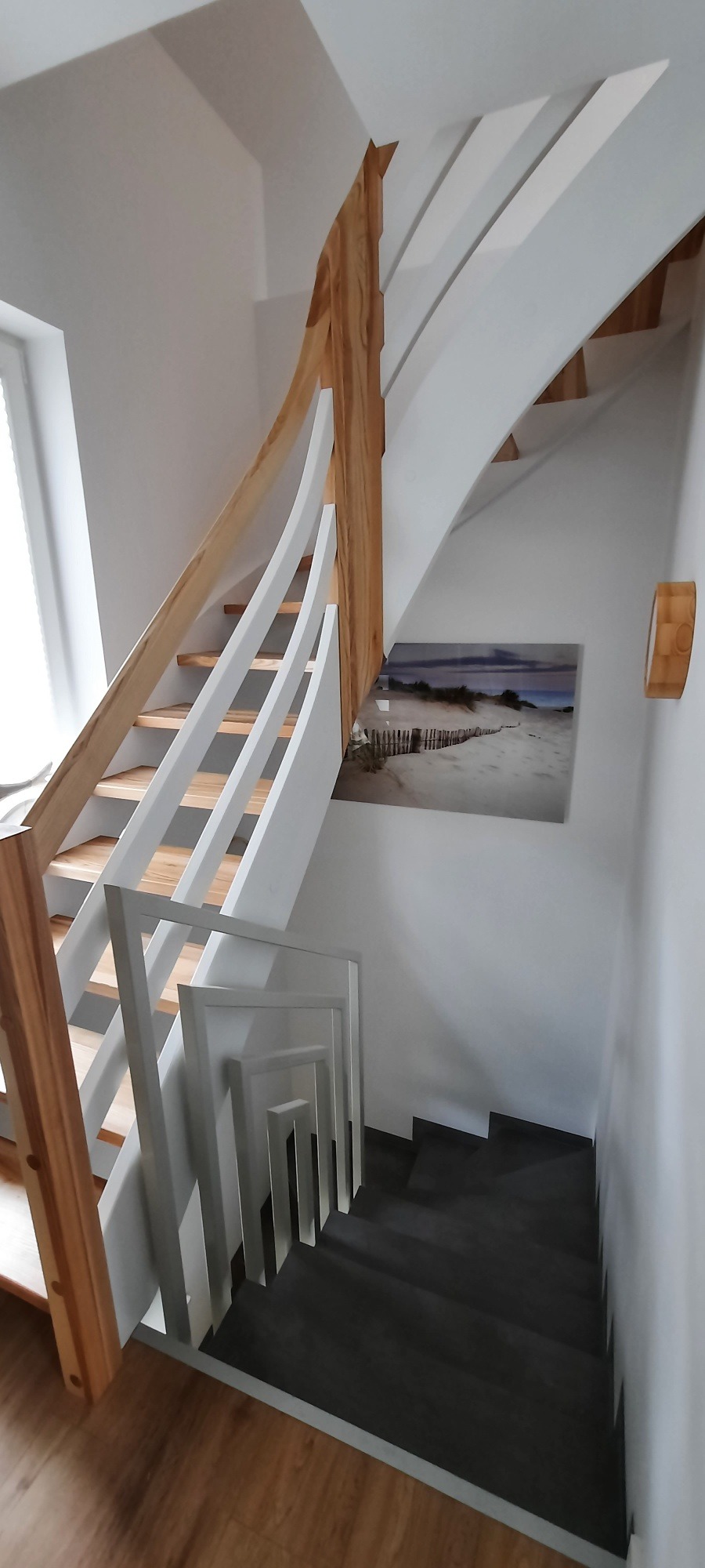 Spiralne schody z jasnego drewna i białymi balustradami, ciemne stopnie, na ścianie obraz z motywem plaży, widok z góry.