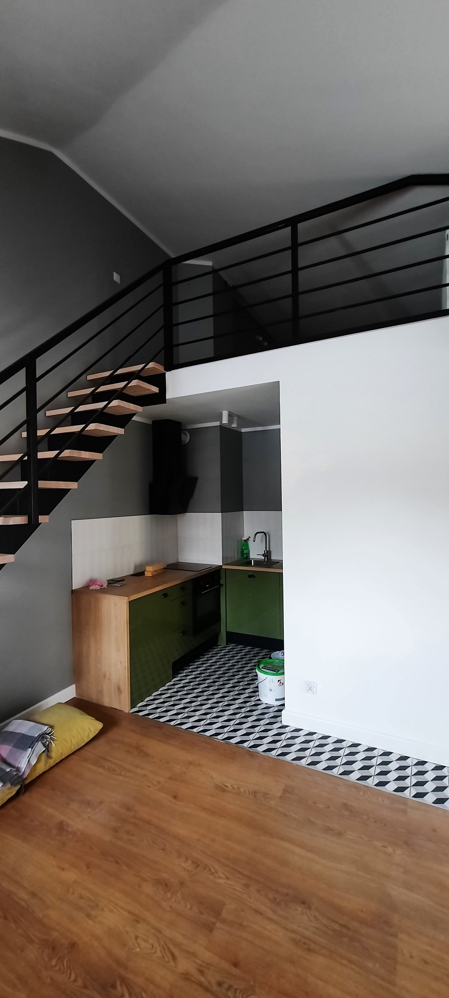 Nowoczesna kuchnia w aneksie z zielonymi szafkami, drewnianym blatem i czarno-białą podłogą w geometryczny wzór, widoczna spod drewnianych schodów z czarną balustradą prowadzących na antresolę.