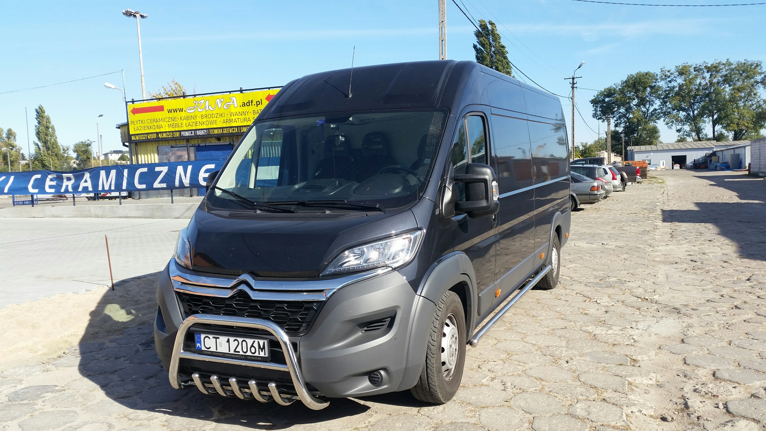 Czarny bus Citroen z relingami i orurowaniem na tle placu z kostki brukowej i budynków. Widoczny szyld reklamowy sklepu z ceramiką.