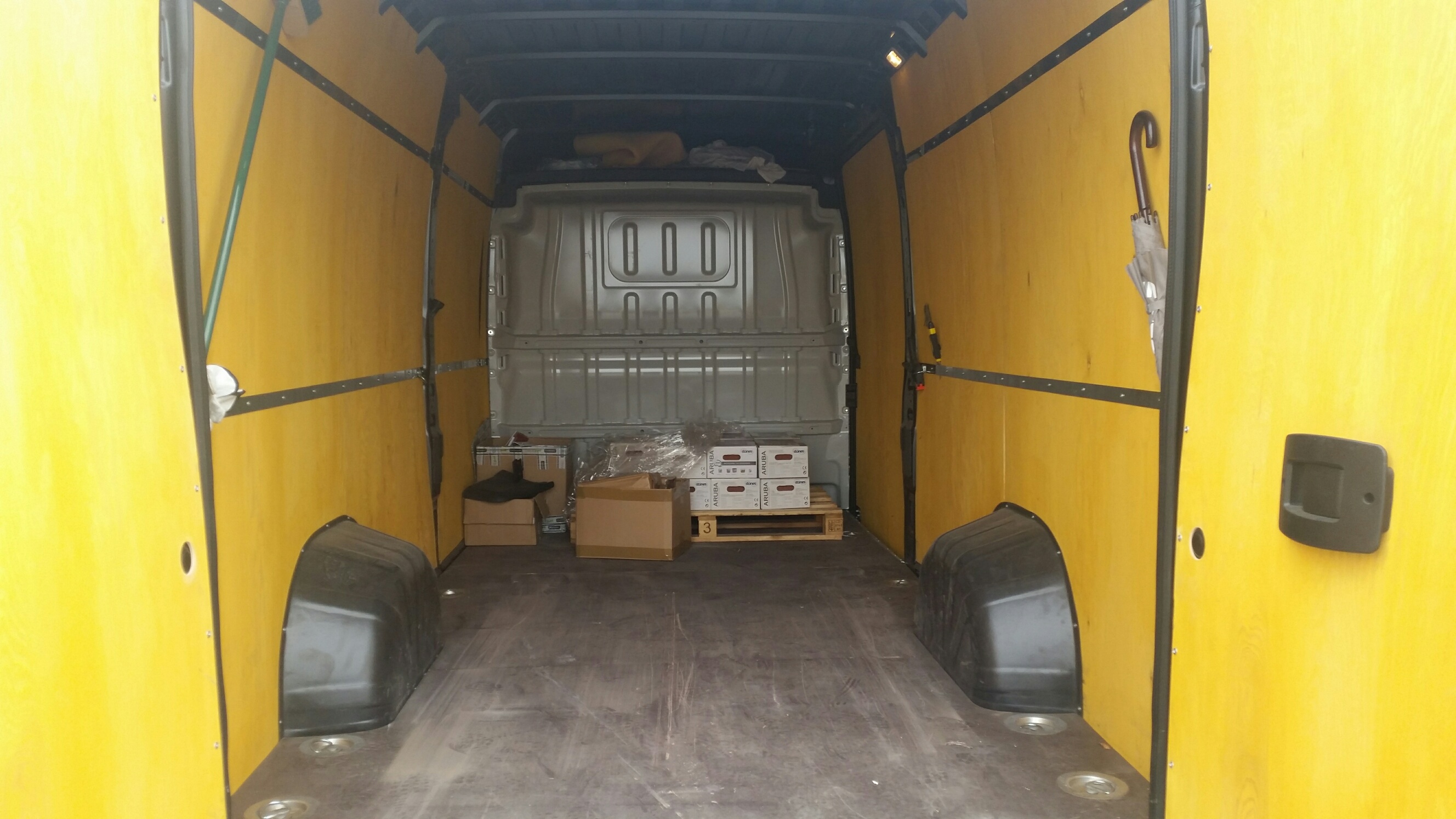 Wnętrze przestrzeni ładunkowej busa wyłożonego żółtą sklejką, z zabezpieczonym ładunkiem w kartonach na palecie. Widoczny parasol po prawej stronie.