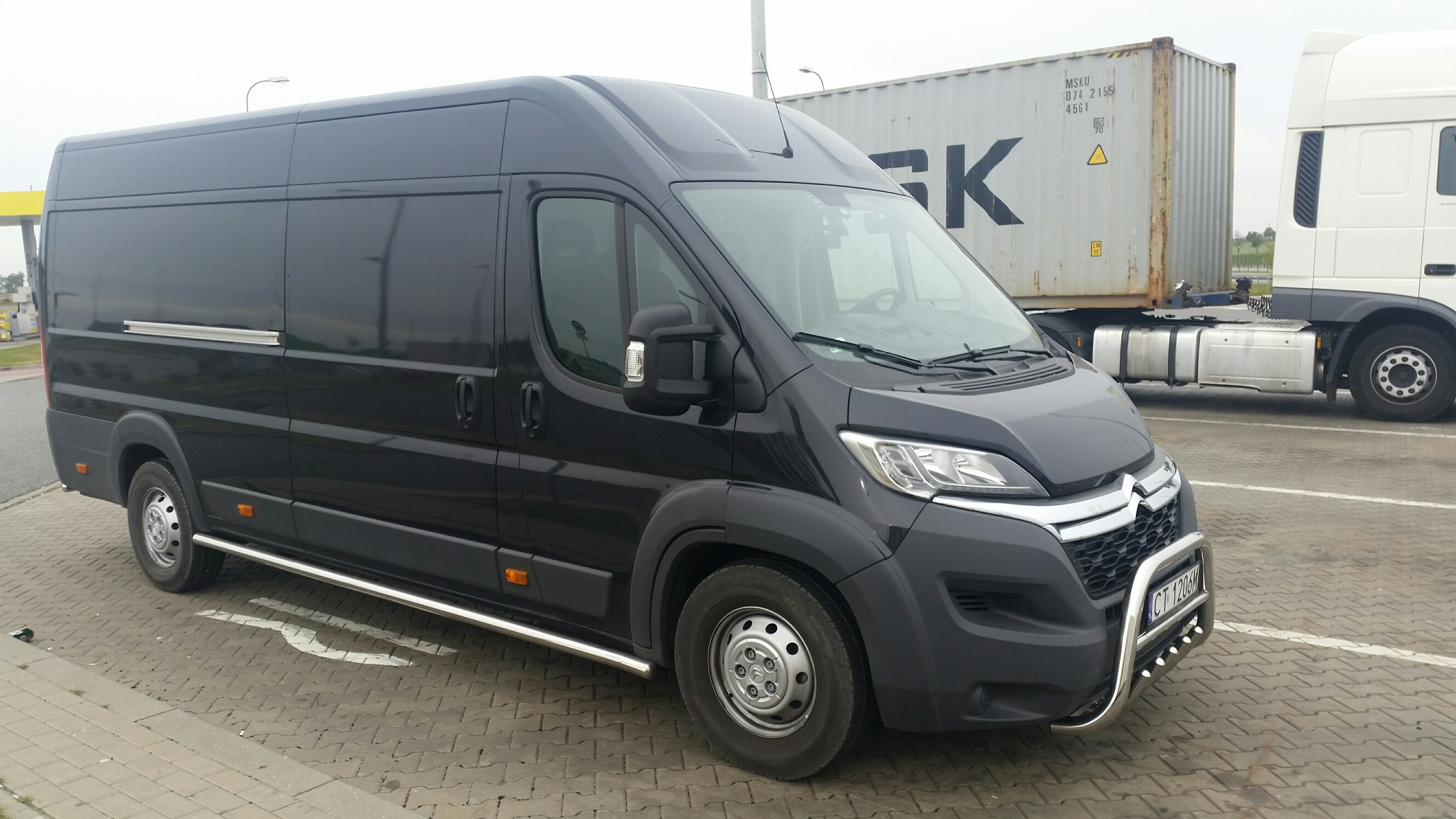 Czarny bus dostawczy z orurowaniem ochronnym na parkingu, obok naczepa z kontenerem. Usługi transportowe, przeprowadzki.