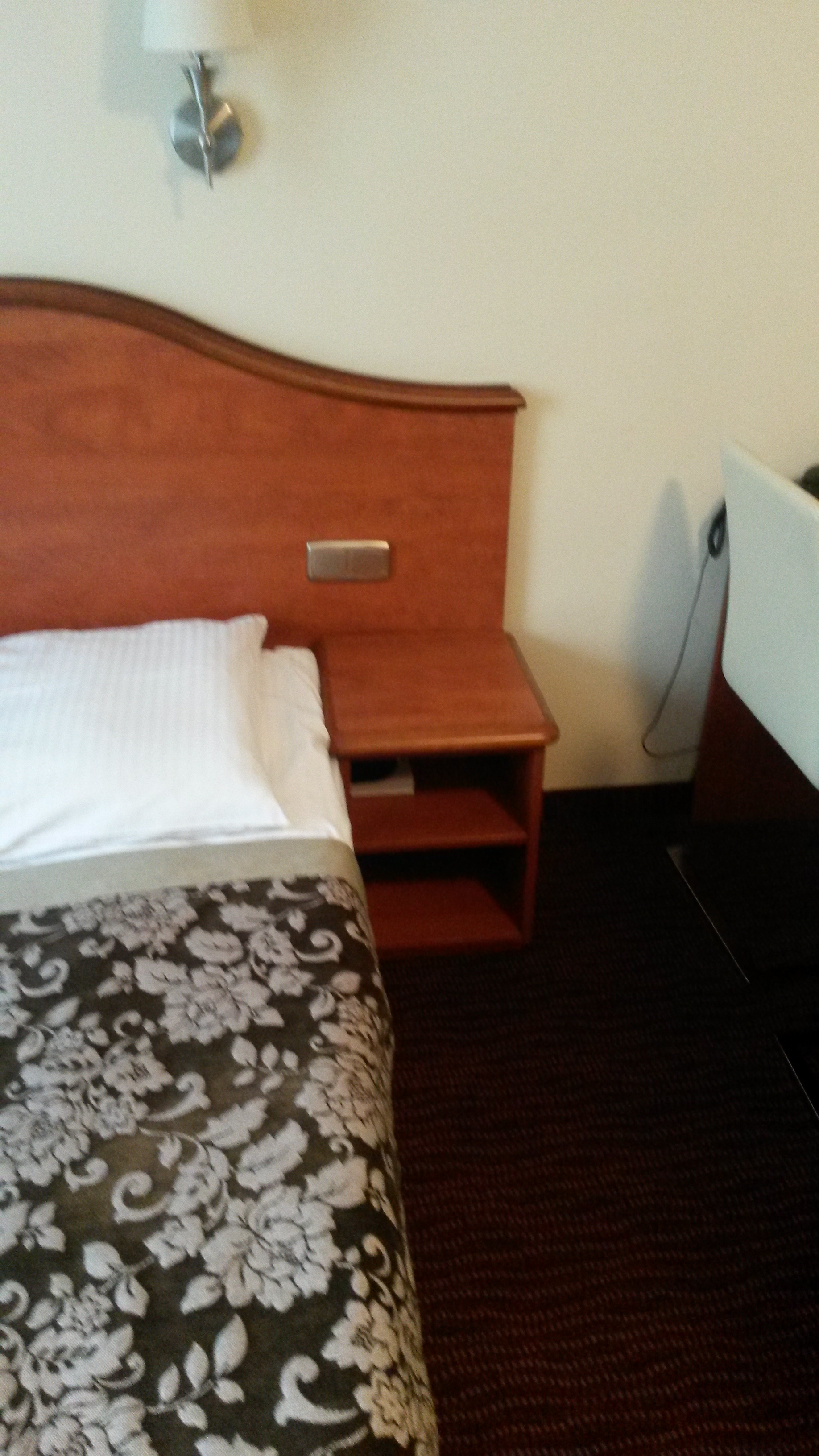 Drewniane wezgłowie z szafką nocną w pokoju hotelowym, widok z perspektywy łóżka. Na ścianie kinkiet, obok biurko z krzesłem. Ciemna wykładzina podłogowa.