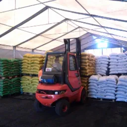 Czerwony wózek widłowy Linde na tle palet z workami z opałem w magazynie z zadaszeniem z plandeki.