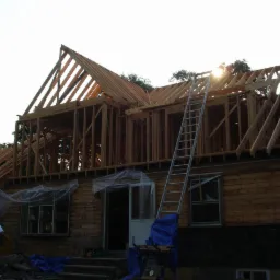 Konstrukcja domu szkieletowego w trakcie budowy: drewniane belki, ściany i dach. Długa drabina oparta o budynek. Widoczny pracownik i zabezpieczenia okien folią.