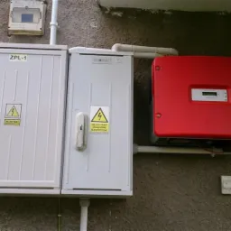 Zewnętrzna instalacja elektryczna: dwie skrzynki z ostrzeżeniami i czerwony inwerter SMA na szarej elewacji budynku z numerem 268.
