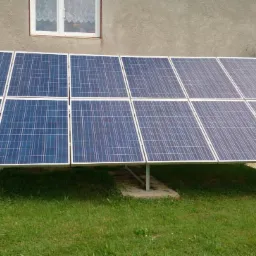 Instalacja fotowoltaiczna na trawniku przed budynkiem z oknem i antenami satelitarnymi. Panele ustawione na metalowych wspornikach.