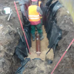 Montaż rury w wykopie zabezpieczonym folią, dwóch pracowników w odblaskowych kamizelkach, różowy kask, koparka w tle. Prace ziemne przyłącza wod-kan.