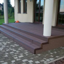 Trzy stopniowe wejście do domu wykończone brązowym tarasem kompozytowym, z widoczną strukturą desek, kolumna podtrzymująca zadaszenie, kostka betonowa na podjeździe, w tle domek dla dzieci w ogrodzie.