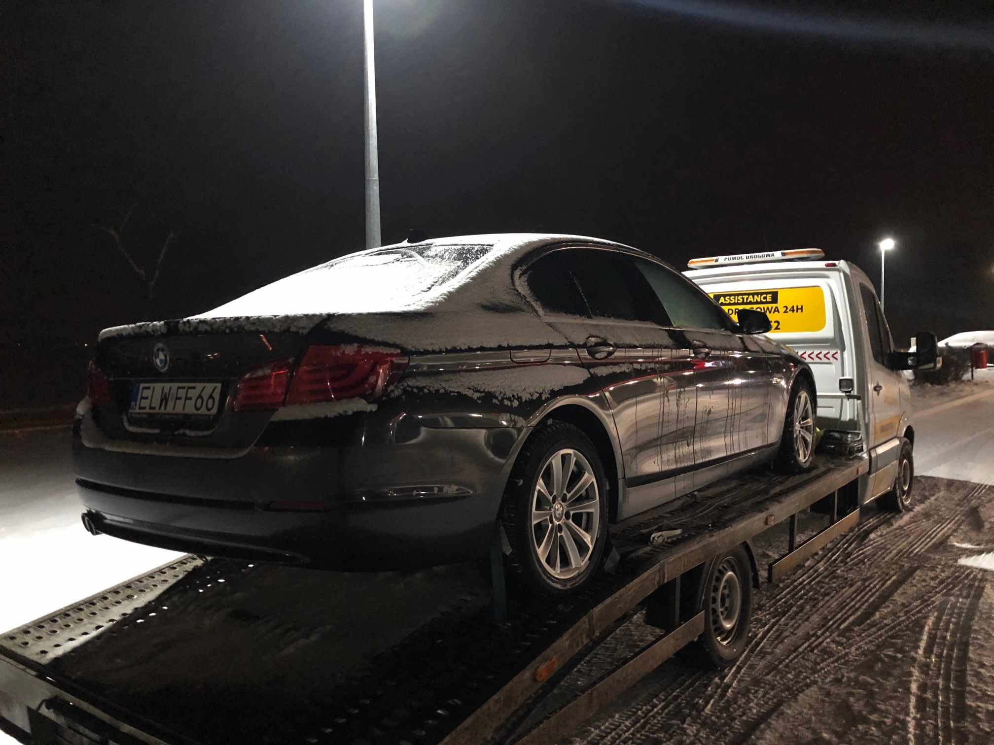Ciemny samochód BMW z tablicą rejestracyjną ELW FF66, lekko pokryty śniegiem, umieszczony na platformie lawety w nocnej scenerii.