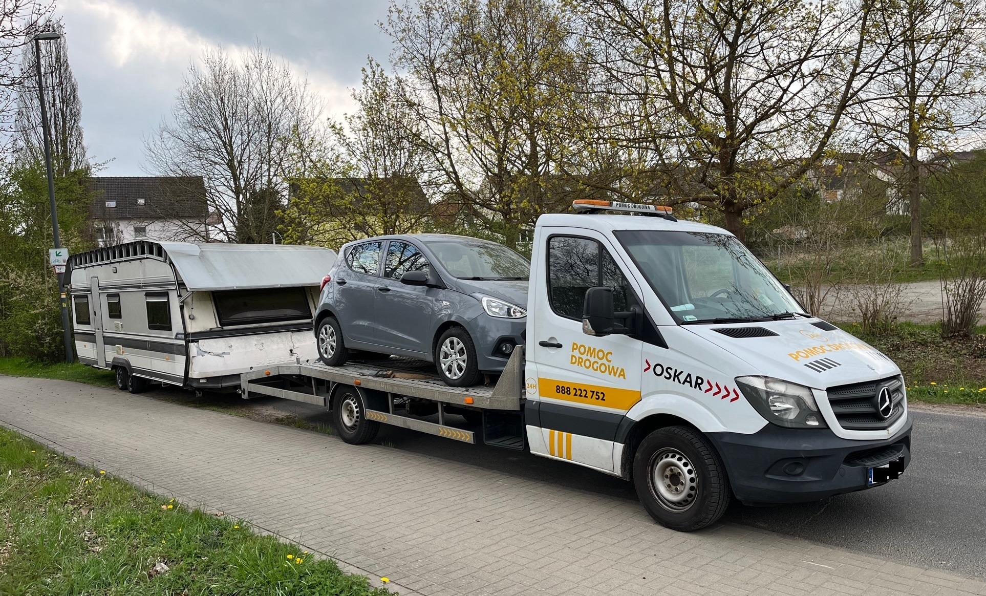 Laweta z szarym samochodem osobowym i przyczepą kempingową zaparkowana na chodniku, widoczny napis 'POMOC DROGOWA OSKAR' na białej kabinie pojazdu.