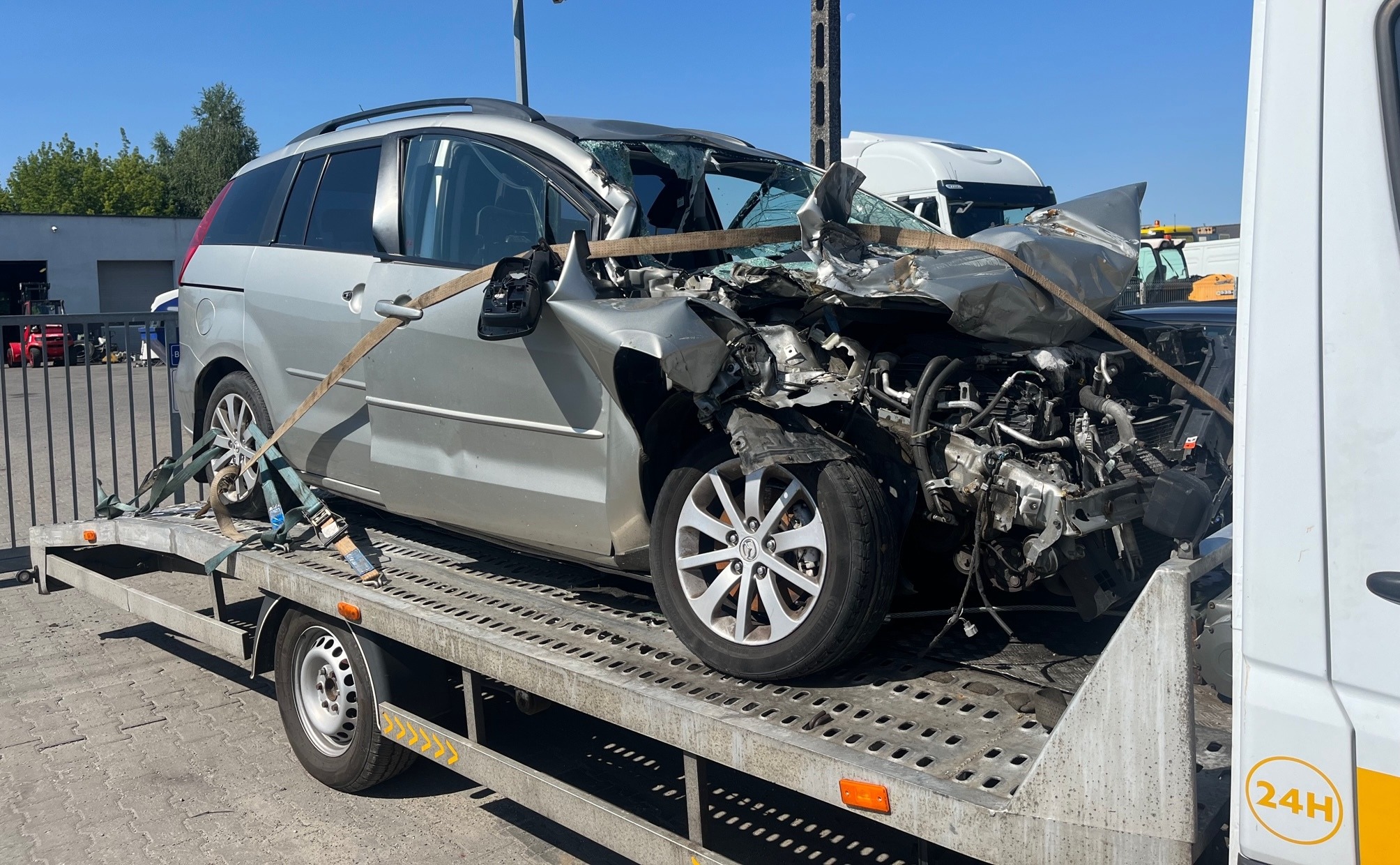 Srebrny, mocno uszkodzony samochód osobowy marki Mazda, zabezpieczony pasami transportowymi, przewożony na lawecie z ażurowaną platformą, widoczne zniszczenia przedniej części pojazdu i rozbite...