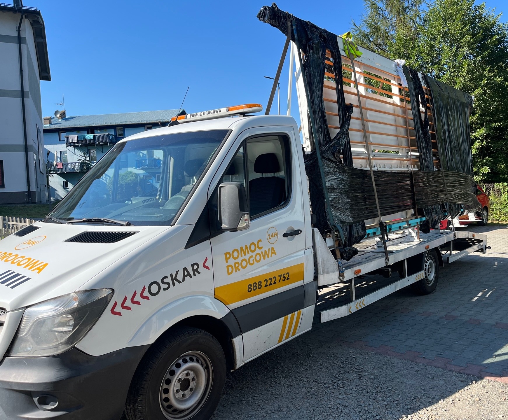 Laweta z Kalwarii Zebrzydowskiej przewozi zabezpieczoną folią konstrukcję, prawdopodobnie okna lub elementy szklane. Na białej kabinie pojazdu widnieje napis 'Pomoc Drogowa' i numer telefonu.