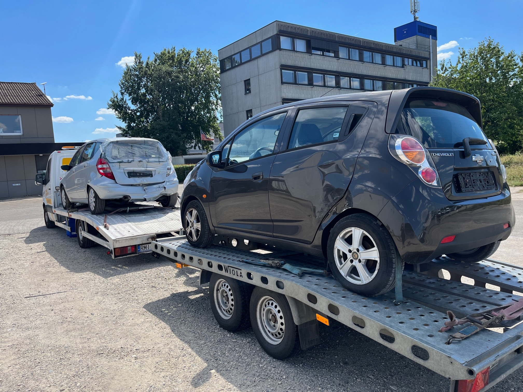 Dwa uszkodzone samochody, srebrny Mercedes z zaklejoną tylną szybą i ciemnoszary Chevrolet Spark z wgnieceniami, przewożone na platformie lawety w słoneczny dzień.