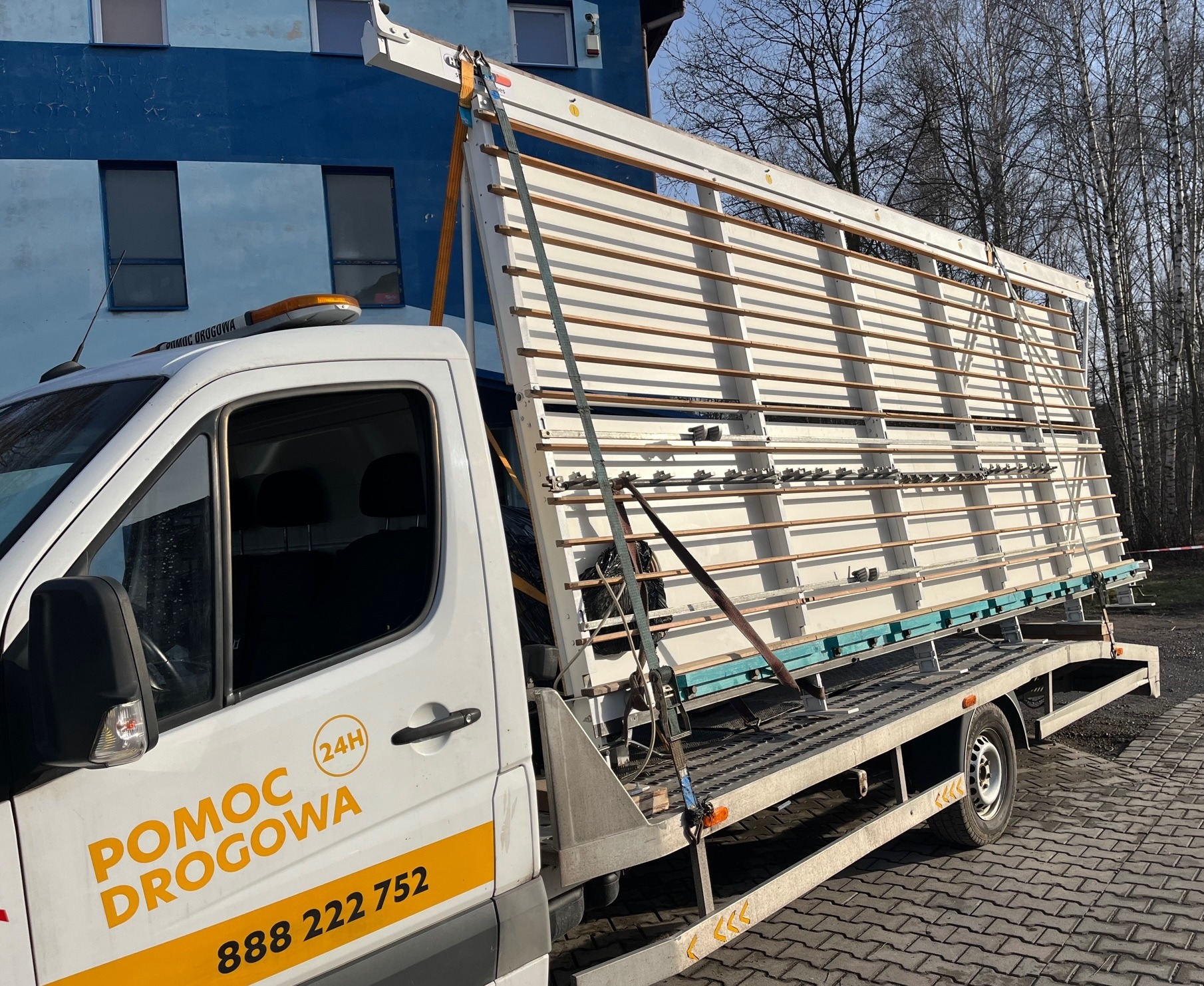 Laweta z białym samochodem pomocy drogowej przewozi pionowo ustawione szklane tafle w specjalnej ramie zabezpieczonej pasami transportowymi na tle budynku i drzew.