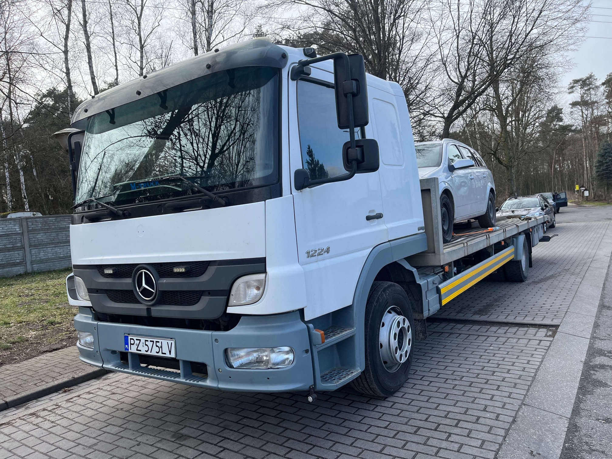 Biała laweta Mercedes-Benz 1224 z załadowanym srebrnym kombi Opel Astra na tle brukowanej drogi i drzew bez liści.