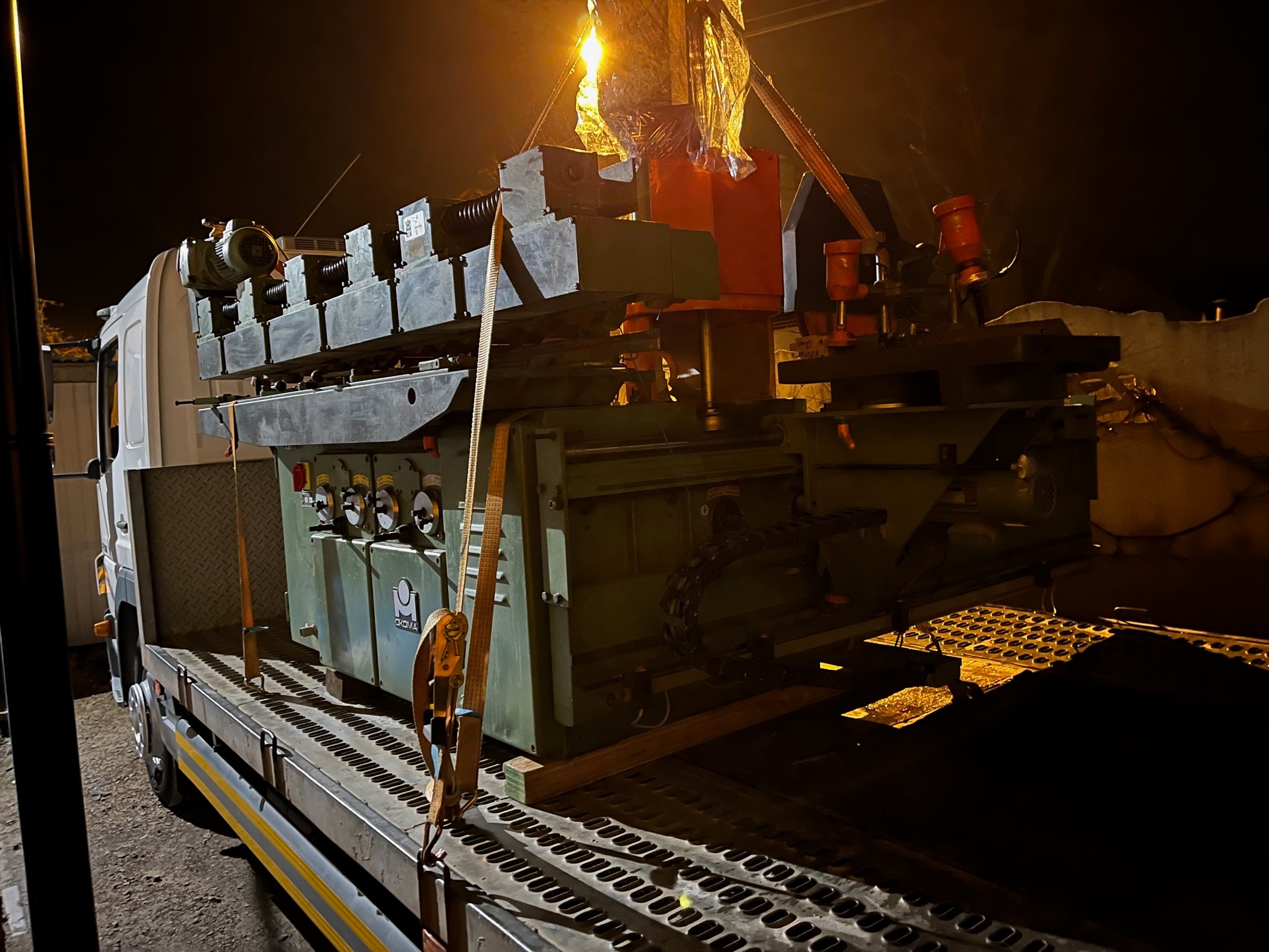 Ciężka maszyna przemysłowa, zabezpieczona pasami transportowymi, przewożona na platformie ciężarówki w nocy.