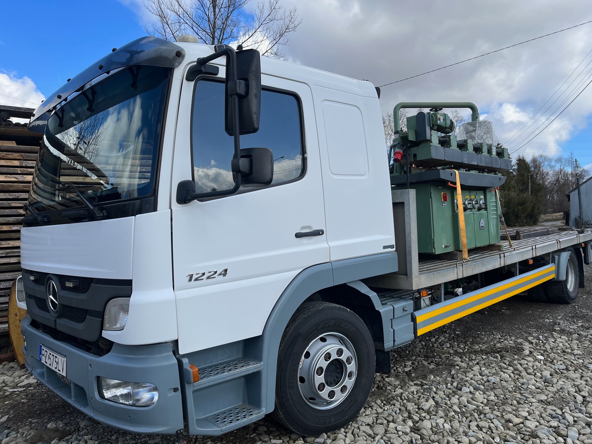 Biała ciężarówka Mercedes-Benz 1224 z platformą załadunkową przewożąca zieloną maszynę przemysłową zabezpieczoną pasami.