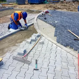 Pracownik w niebieskim kombinezonie i pomarańczowej kamizelce układa szare kostki brukowe na przygotowanym podłożu, część powierzchni wyłożona ciemnoszarą kostką, widoczne narzędzia pracy...