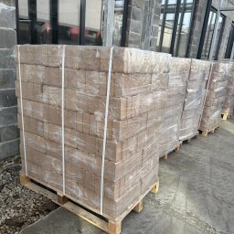 AMT Anthony Vandeginste - Ułożone na paletach, owinięte folią pakiety brykietu drzewnego przed budynkiem z szarej cegły i dużymi oknami.