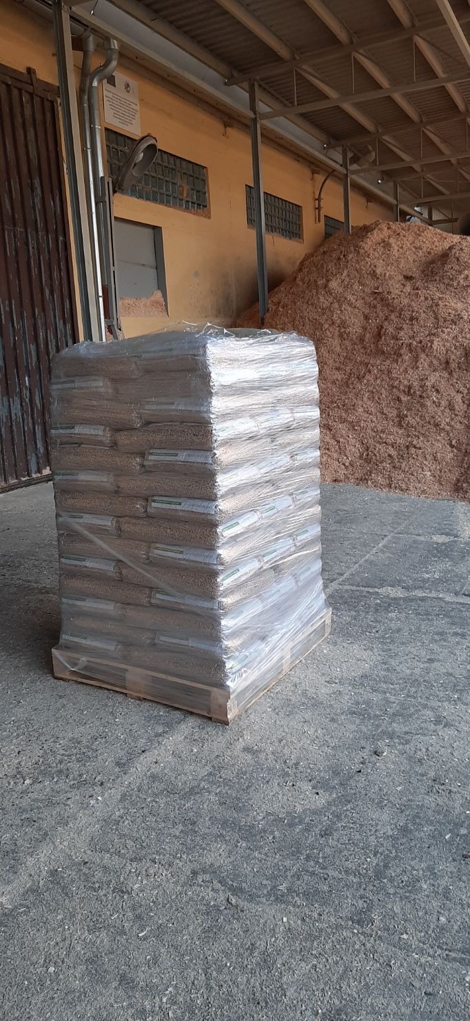 Paleta z workami z granulatem, owinięta folią, na tle hałdy trocin w magazynie.