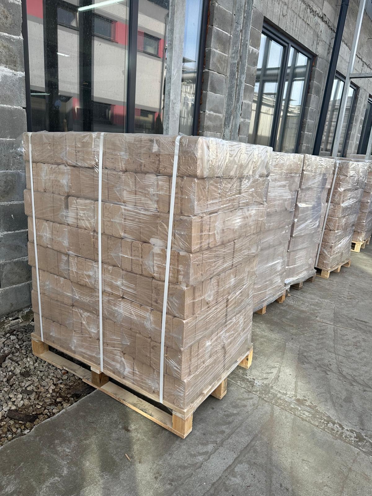 Ułożone na paletach, owinięte folią pakiety brykietu drzewnego przed budynkiem z szarej cegły i dużymi oknami.