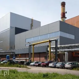 Nowoczesny budynek przemysłowy z szarymi płytami warstwowymi i rurami w tle, z zaparkowanymi autami i zielenią na pierwszym planie.