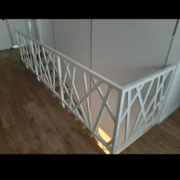 Biała barierka z geometrycznym wzorem na schodach, widok z góry na drewnianą podłogę i białe ściany.