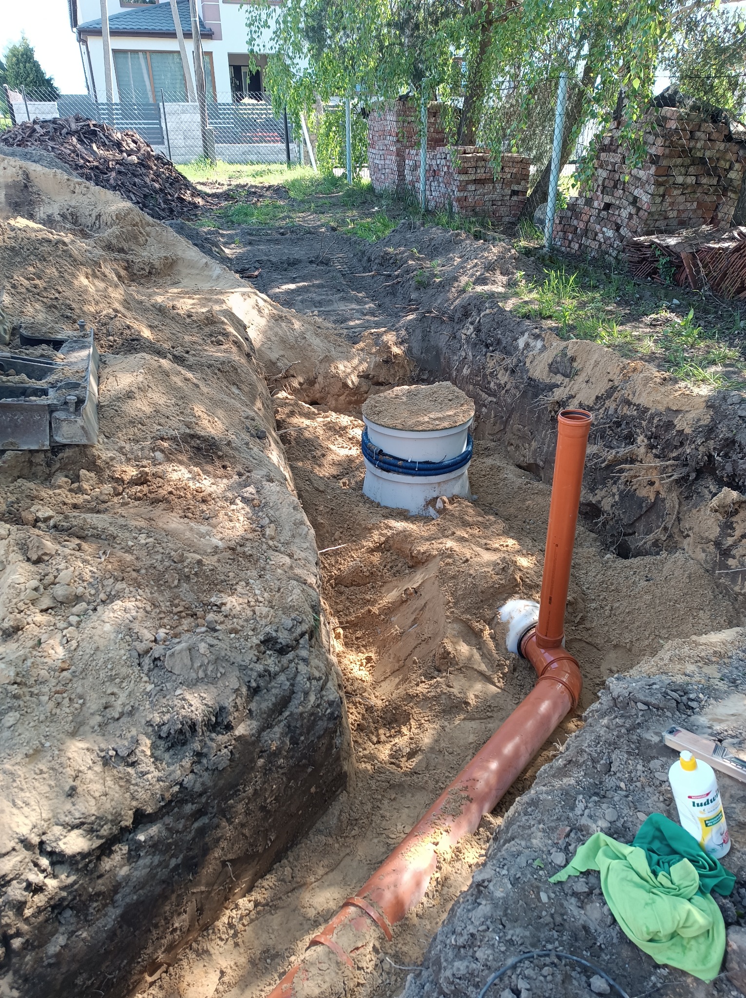 Wykop w ziemi z widoczną studzienką kanalizacyjną z szarym deklem oraz pomarańczowymi rurami PVC, obok leży butelka płynu do mycia naczyń i zielona ścierka.