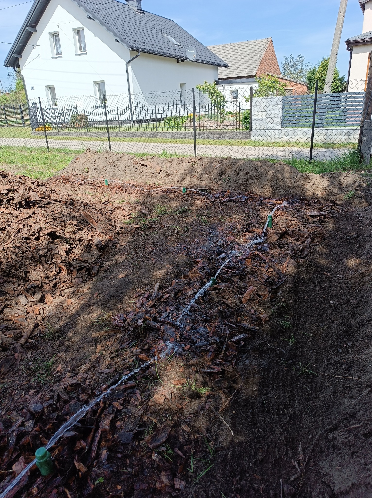 Instalacja nawadniania w ogrodzie z widocznymi rurami i dyszami rozprowadzającymi wodę na korę i ziemię, na tle domu jednorodzinnego.