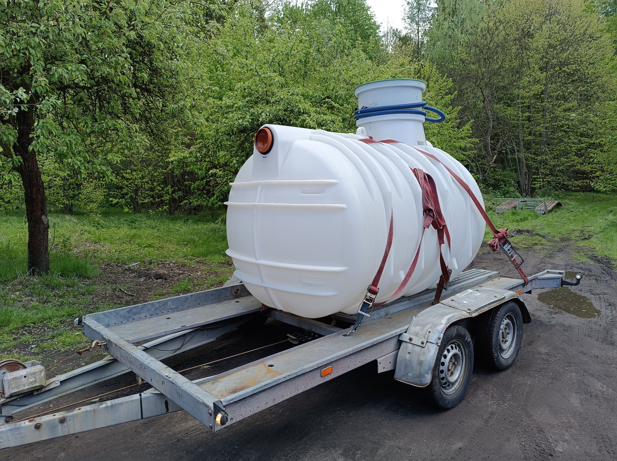 Biały, plastikowy zbiornik na szarej przyczepie transportowej, zabezpieczony czerwonymi pasami, na tle zieleni drzew.
