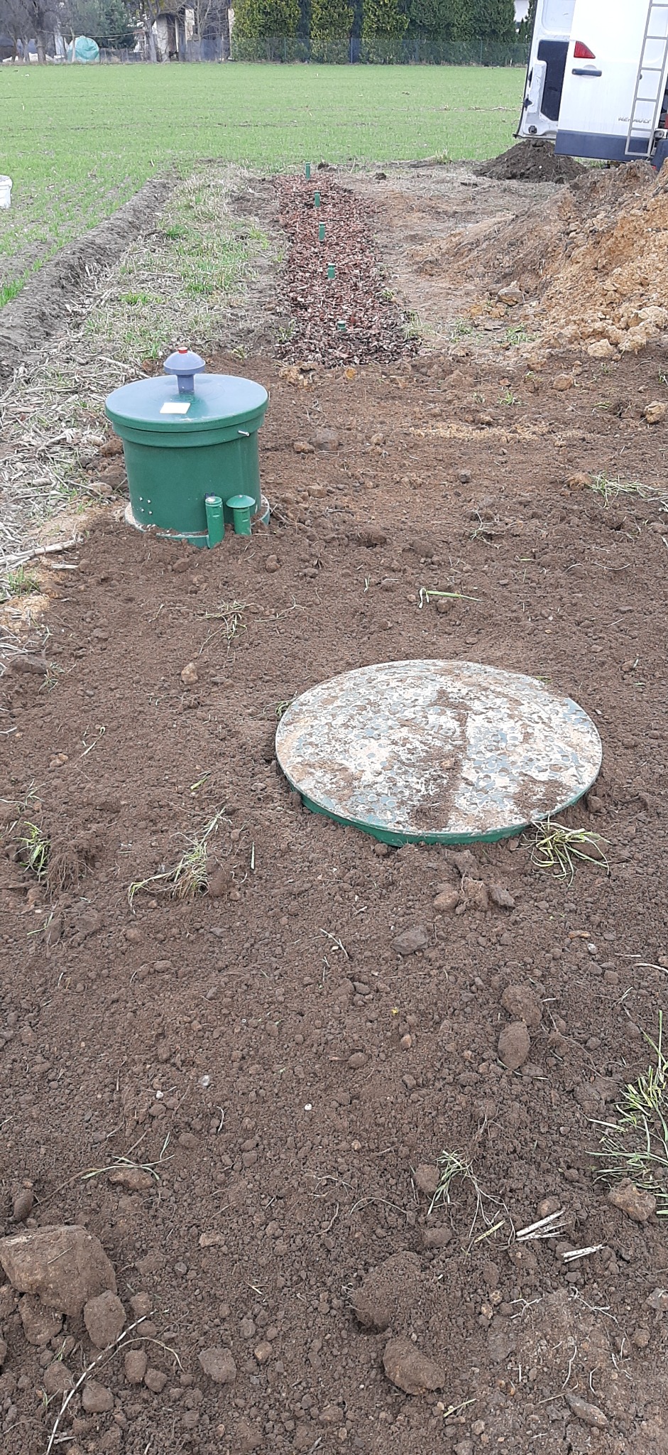 Zielony zbiornik i przykrywa oczyszczalni ścieków na tle świeżo przekopanej ziemi, z widocznym polem uprawnym w tle.