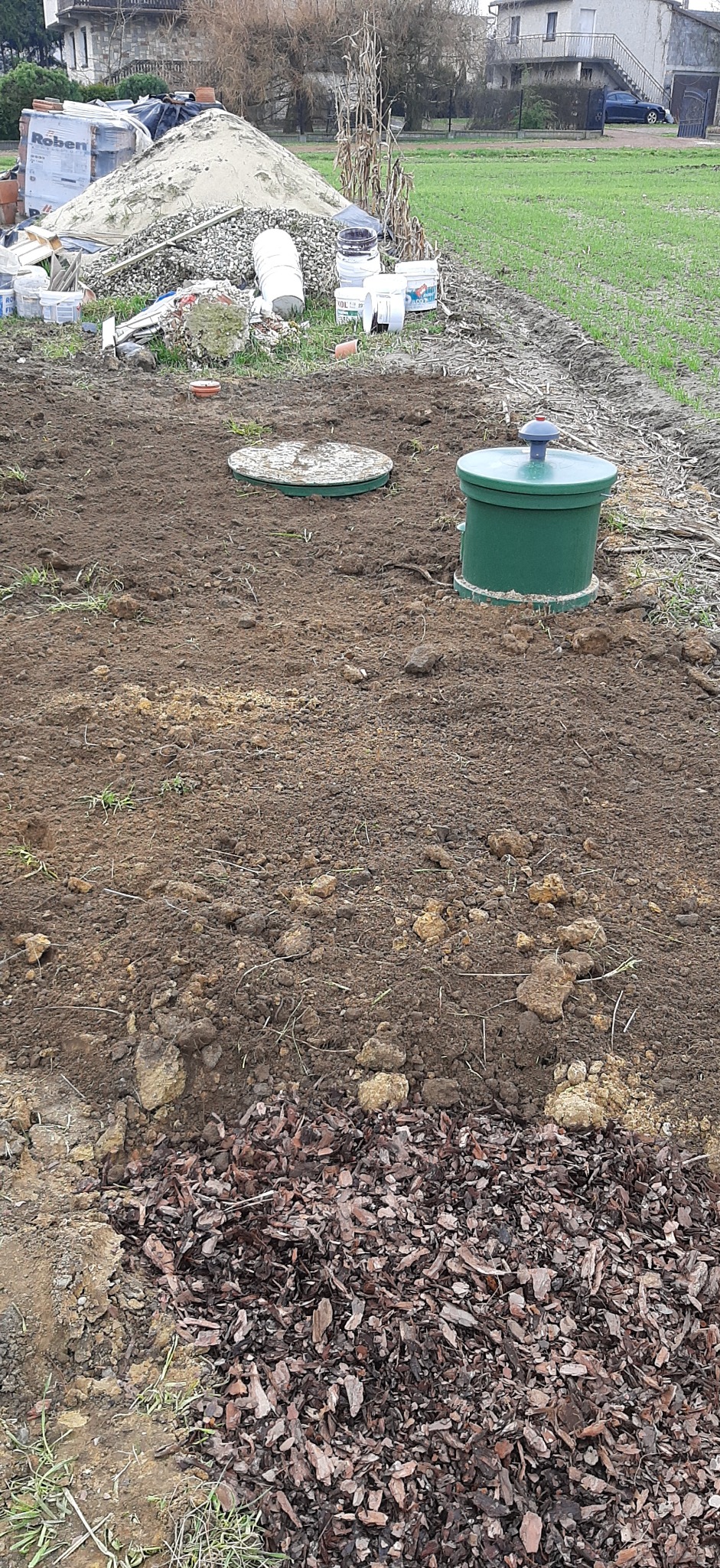 Instalacja przydomowej oczyszczalni ścieków: widoczny zielony zbiornik z szarą pokrywą oraz betonowa pokrywa studzienki na przygotowanym terenie, w tle sterta piasku i materiałów budowlanych.