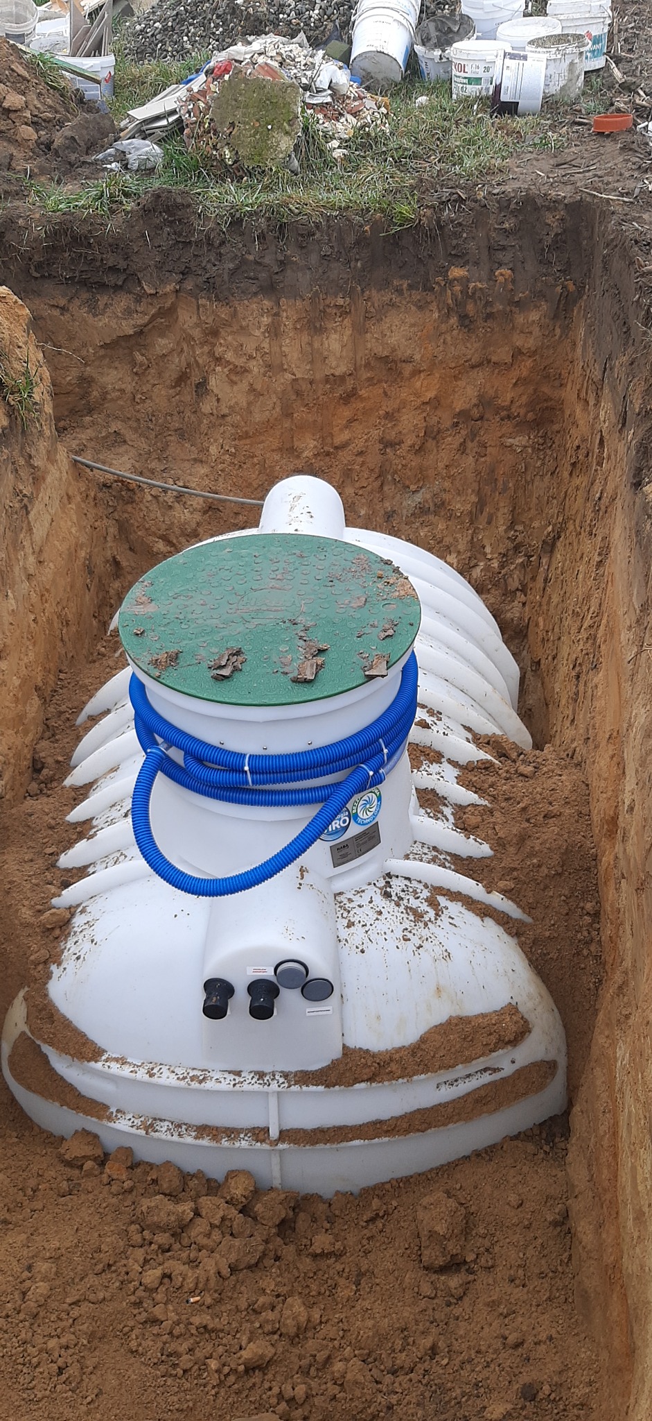 Montaż zbiornika oczyszczalni ścieków w wykopie, widoczne niebieskie rury i zielona pokrywa, ziemia wokół. W tle widoczne materiały budowlane i gruz.