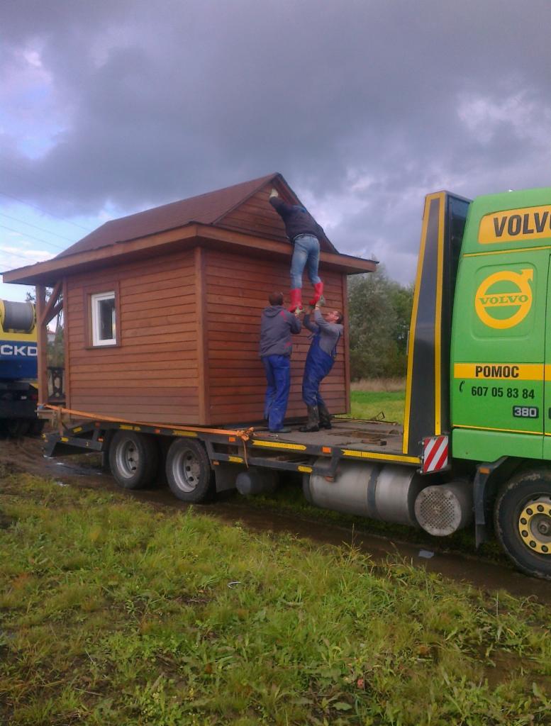Transport domku drewnianego na lawecie Volvo. Trzech mężczyzn zabezpiecza dach podczas transportu. Jeden stoi na ramionach kolegów w czerwonych kaloszach.