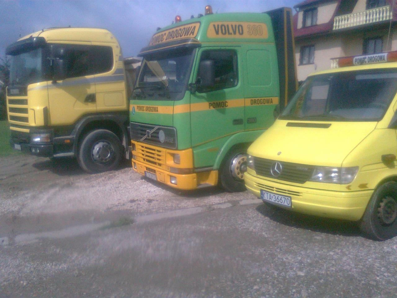 Żółty Scania, zielono-żółty Volvo 380 i żółty Mercedes Sprinter z napisem 'Pomoc Drogowa' na tle budynku.