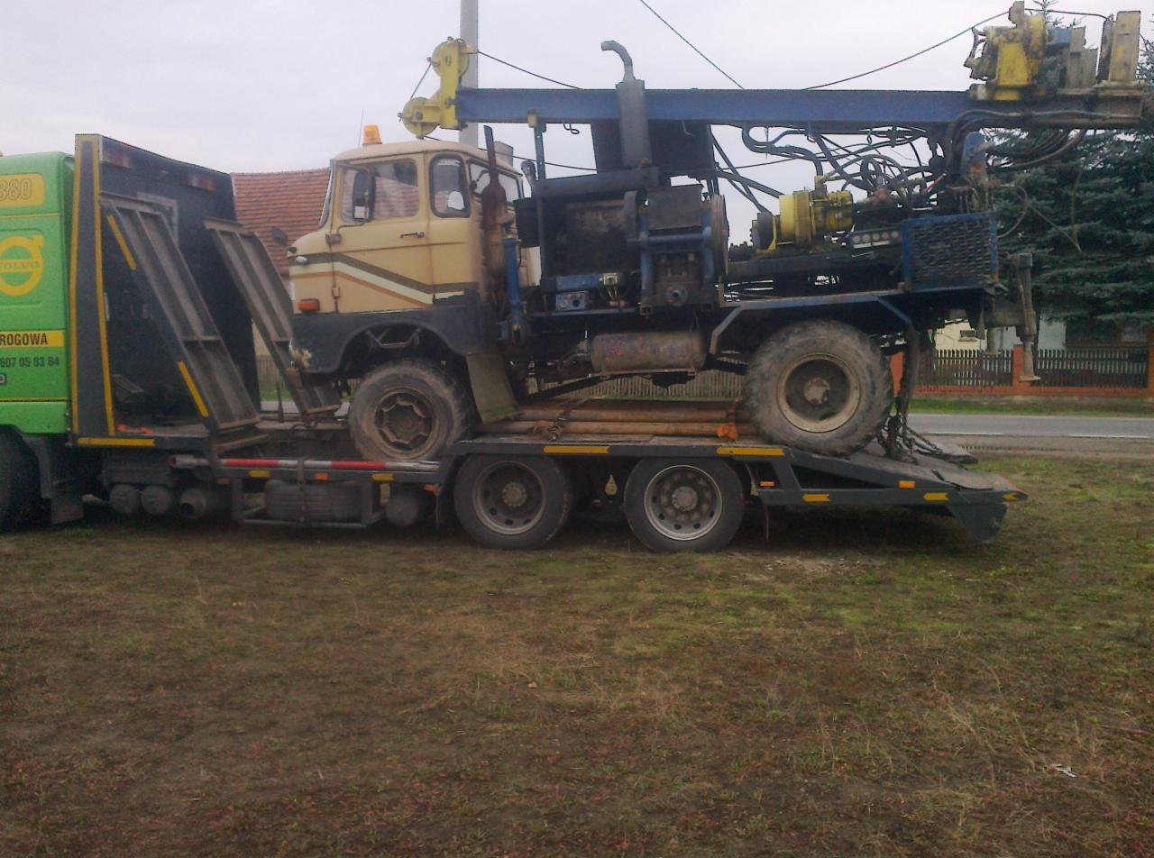 Laweta przewozi wiertnicę na platformie. Widoczny stary model ciężarówki w kolorze beżowym z elementami niebieskimi. Zielona kabina lawety z boku.